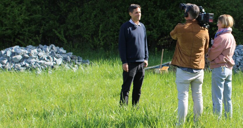 Fachgruppenleiter Holger Schanz bei den Dreharbeiten. Im Hintergrund ist eine Gartenschläfer-Burg zu sehen. Fachgruppenleiter Holger Schanz steht auf einer Wiese. Vor ihm ein Mann, der eine Kamera auf der Schulter trägt und ihn filmt. Neben dem Mann steht die Redakteurin und stellt Fragen. Im Hintergrund ist einer der Steinhaufen für den Gartenschläfer zu sehen.