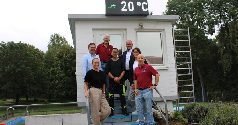 Inna Wedel, Alexander Schaper, Werner Amend, Sascha Schmidt, Bürgermeister Marcus Kretschmann, Dorota Bayram und Holger Michels (v.li.) unter der neuen Schwimmbaduhr. Inna Wedel, Alexander Schaper, Werner Amend, Sascha Schmidt, Bürgermeister Marcus Kretschmann, Dorota Bayram und Holger Michels stehen im Freibad Goddelau auf einer Treppe mit blauen Stufen. Die Treppe führt zu einem weiß gestrichenen Turm. Auf dem Turm befindet sich die neue Uhr. Sie zeigt gerade an "Luft: 20 Grad"